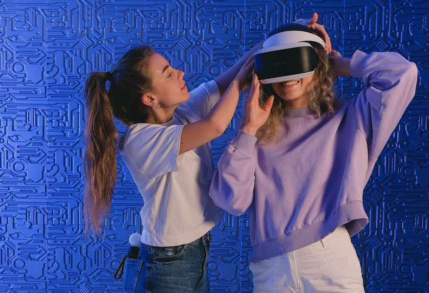 Auf dem Bild sieht man zwei Frauen. Eine trägt eine VR-Brille. Die andere hilft ihr beim Aufsetzen.
